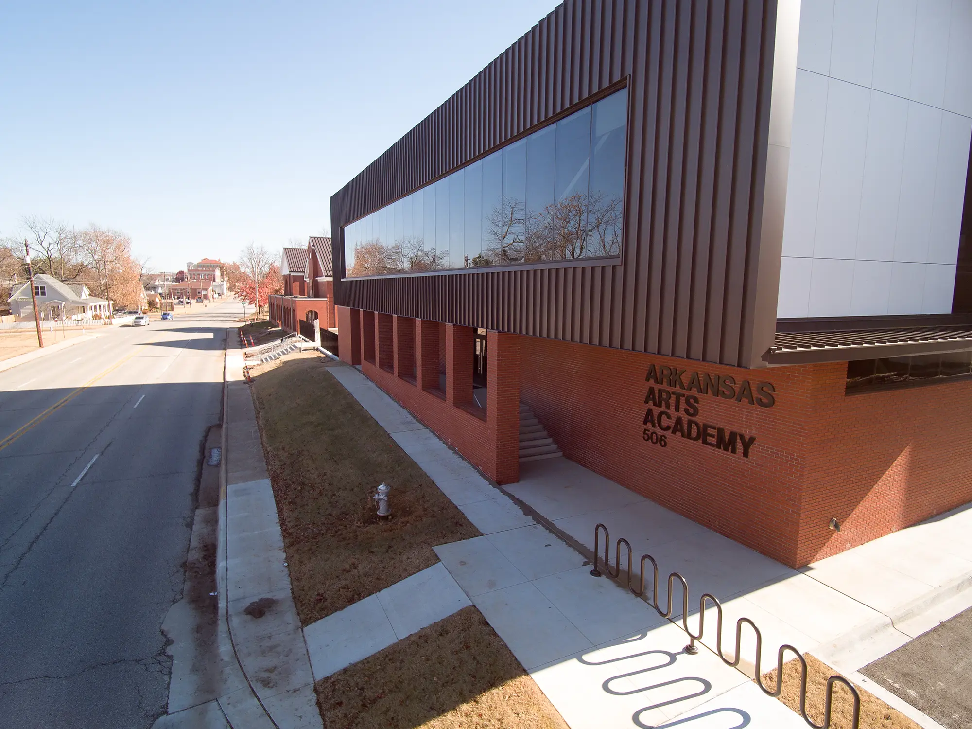 Modern building of Arkansas Arts Academy with red brick and large windows, facing a quiet street with bare trees under a clear blue sky.