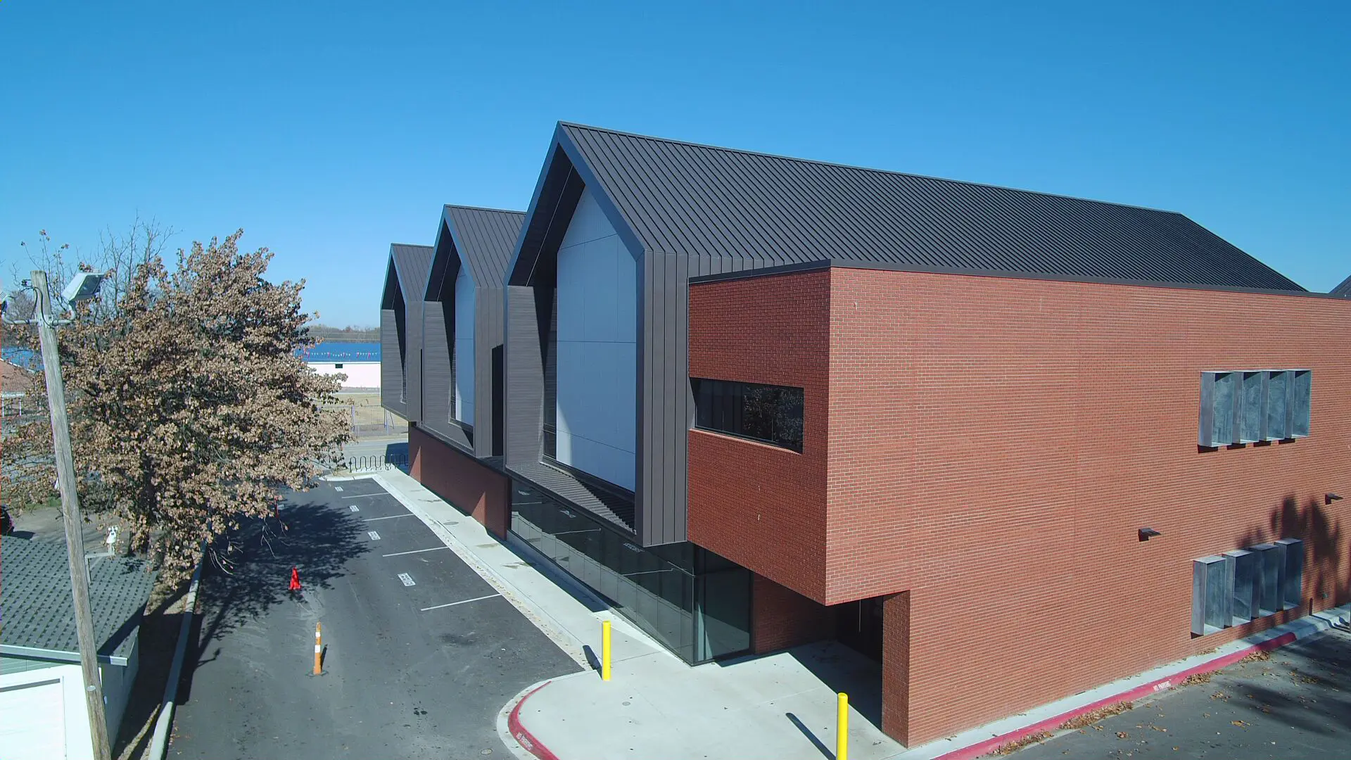 A modern, angular two-story building with a red brick facade and a dark sloped roof against a clear blue sky. Nearby are bare trees and an empty parking area.