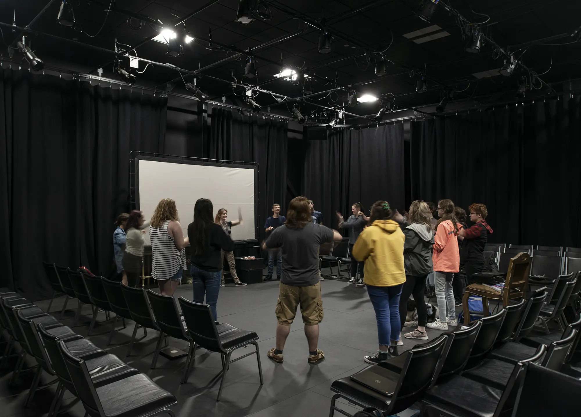 A group of people stand in a circle in a dark room with black curtains, engaging in an activity. The atmosphere is collaborative and focused.