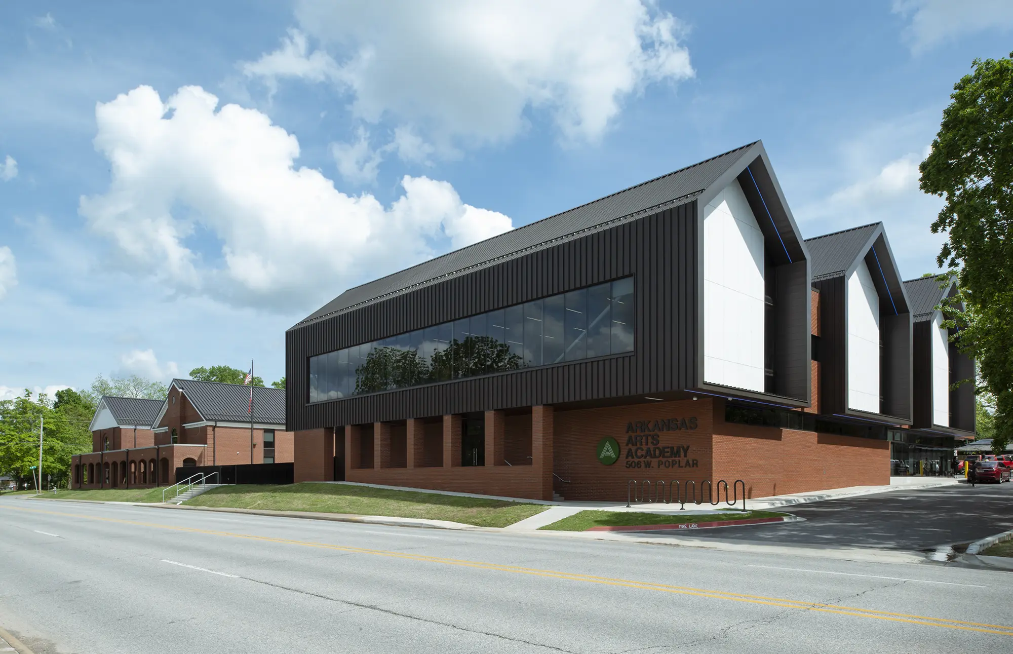 Modern brick and metal building of the Arkansas Arts Academy against a bright blue sky with clouds. The scene is calm and orderly, with nearby trees.