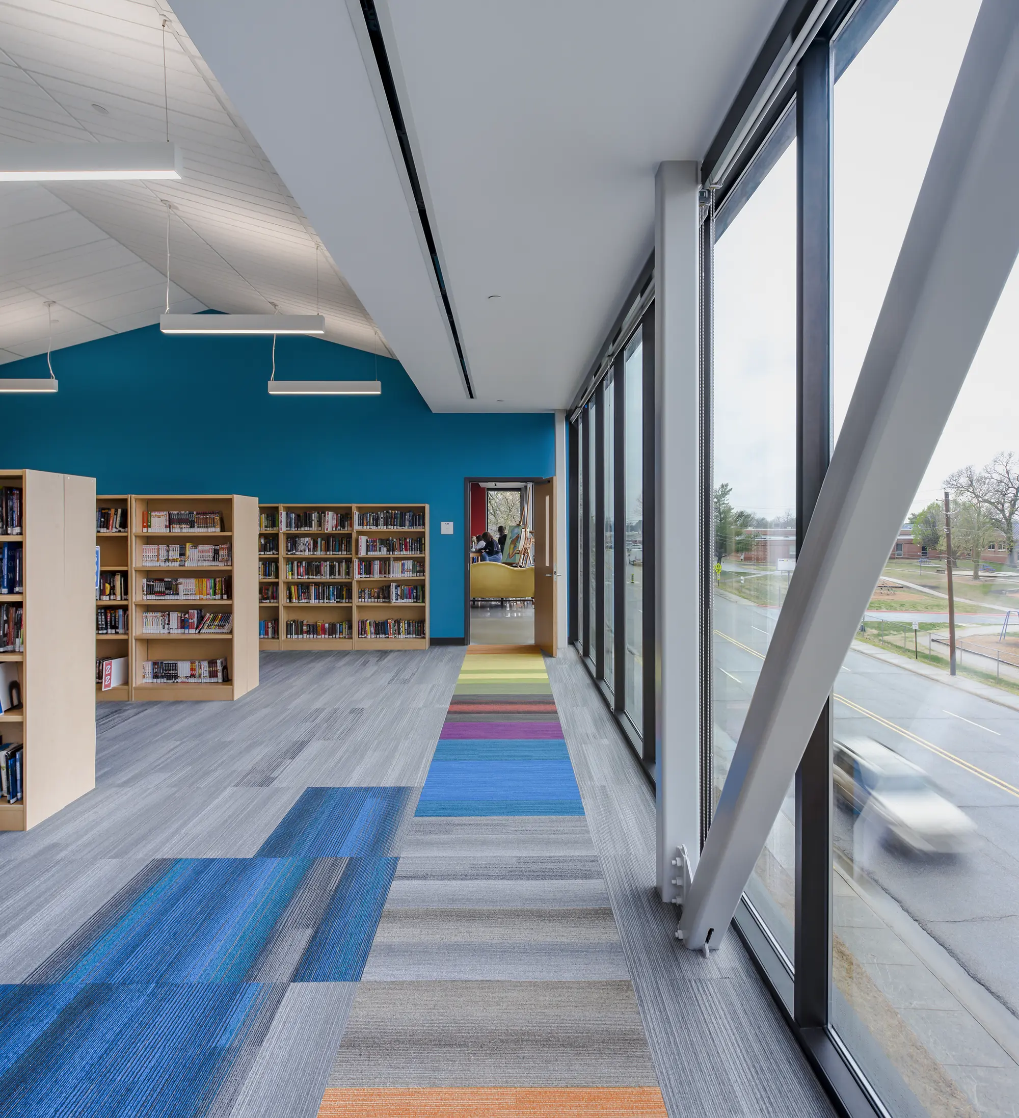 Modern library interior with colorful carpet, wooden bookshelves, and large windows offering a street view. Bright, open, and inviting atmosphere.