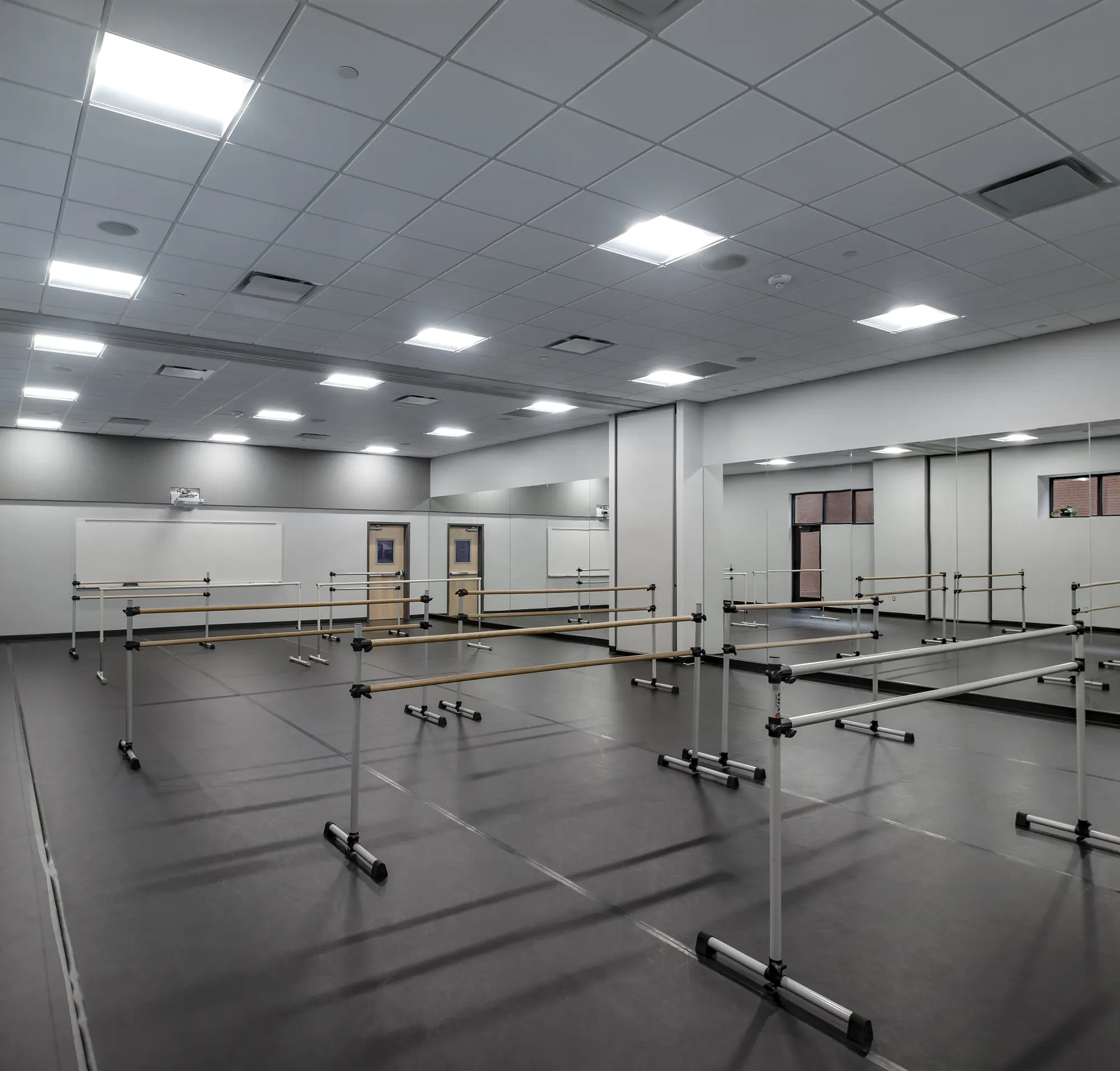 Spacious dance studio with wooden barres, mirrored walls, and gray flooring. Bright overhead lights create a modern, inviting atmosphere for practice.