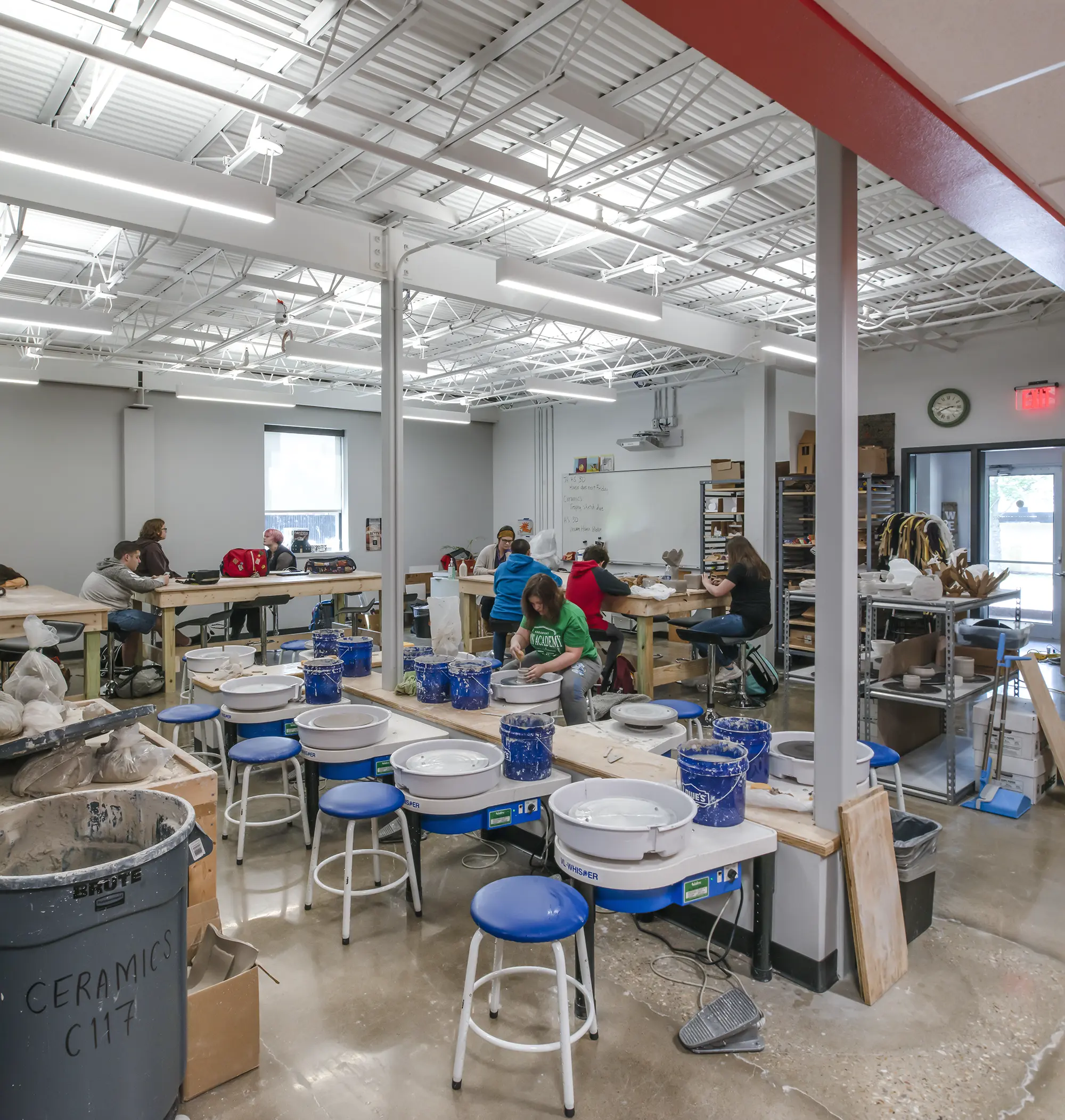 A bright ceramics classroom with students working at tables, surrounded by pottery wheels, clay, and tools. The room has a busy, creative atmosphere.