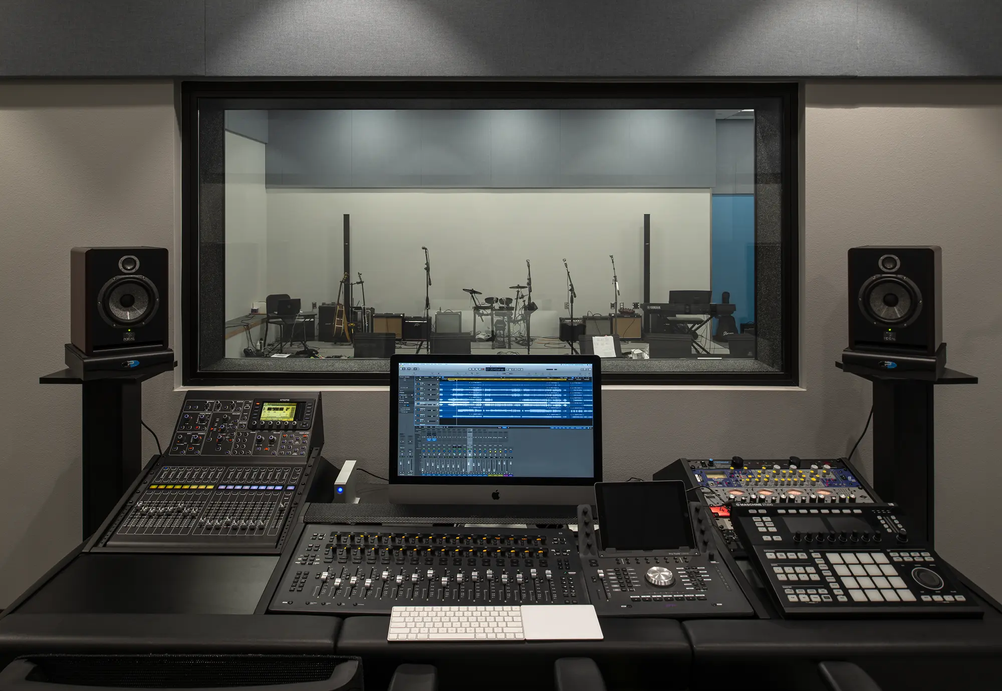 A modern recording studio with a mixing desk and computer screen in the foreground, and a drum set and microphones visible through a large glass window.