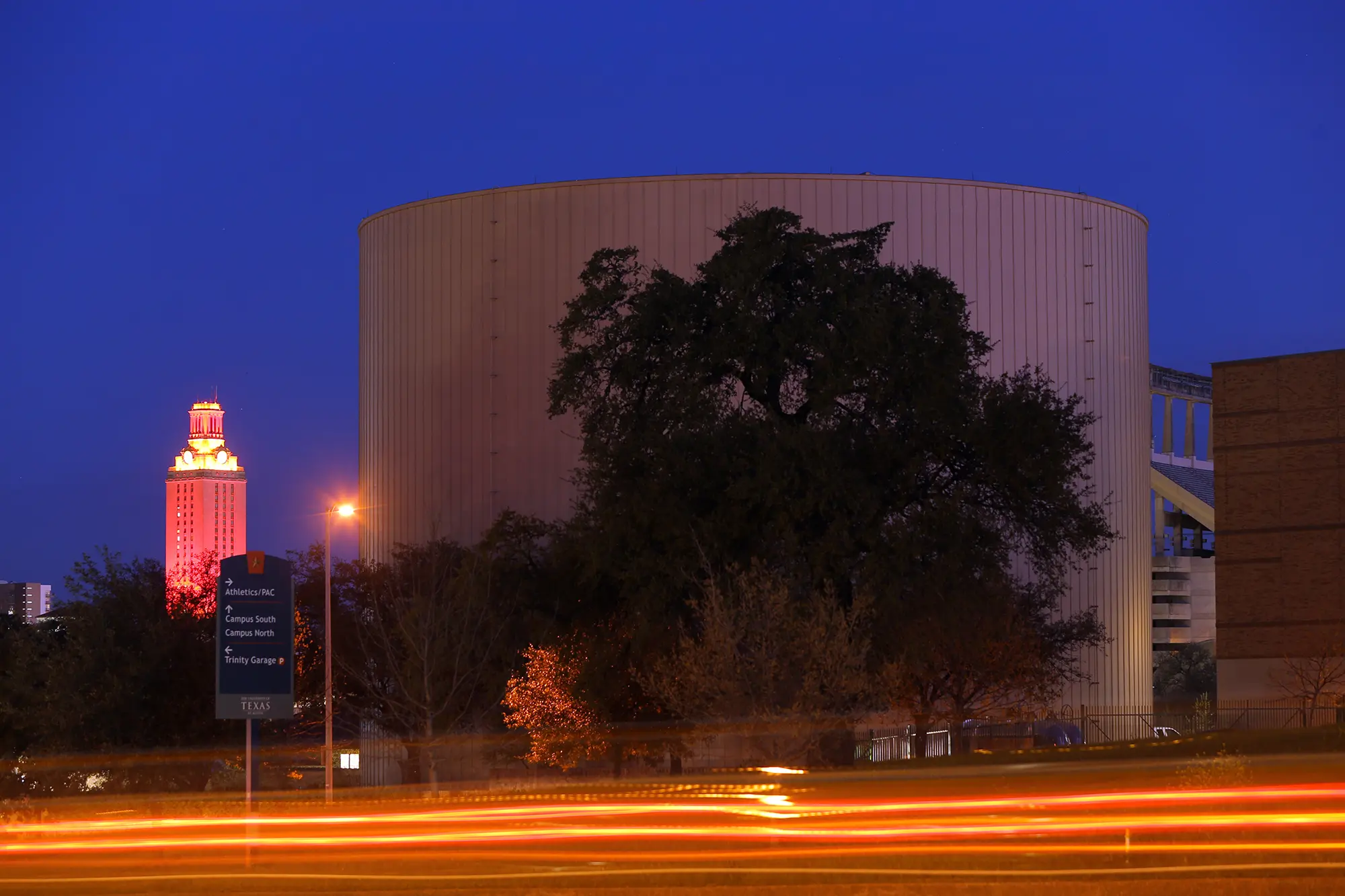 Nighttime cityscape with a tall, glowing orange tower in the distance, surrounded by trees and buildings. Streaks of car lights create motion in the foreground.