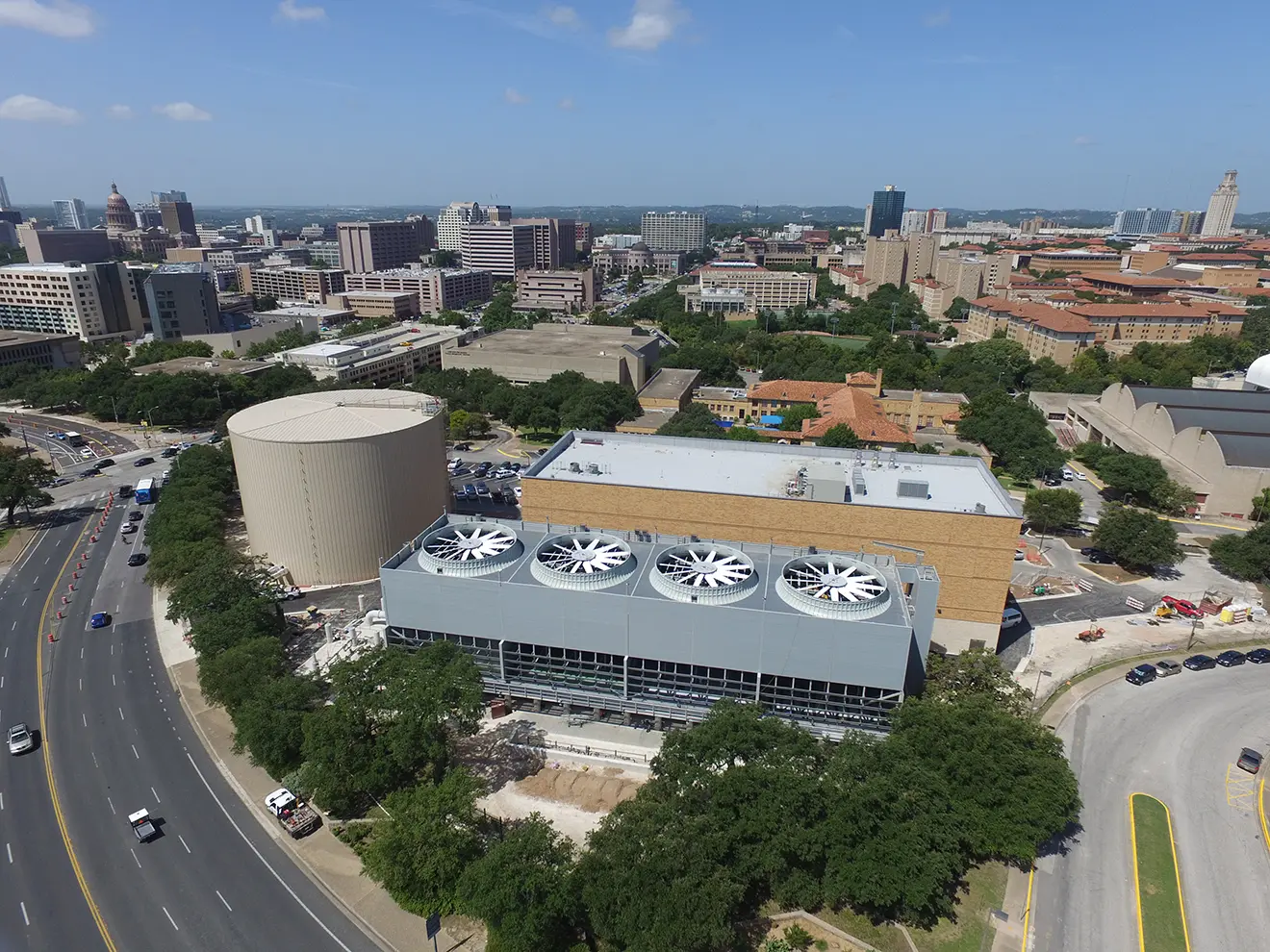 Aerial view of a cityscape featuring a large industrial building with rows of rooftop fans. Surrounding are trees, roads, and high-rise buildings under a clear blue sky.