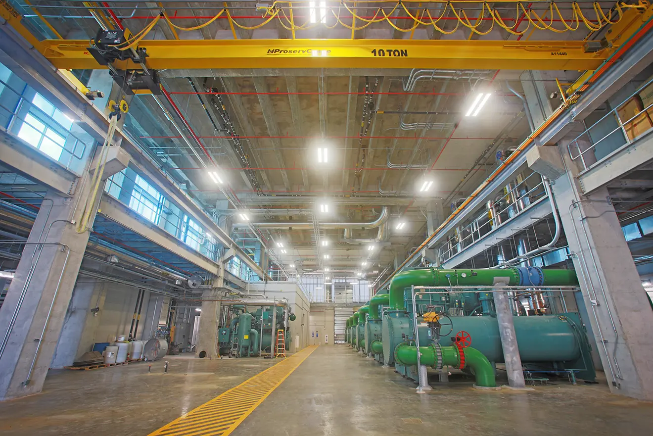 Wide-angle view of an industrial warehouse with high ceilings, featuring large green pipes, a yellow overhead crane labeled "10 TON," and bright overhead lighting.