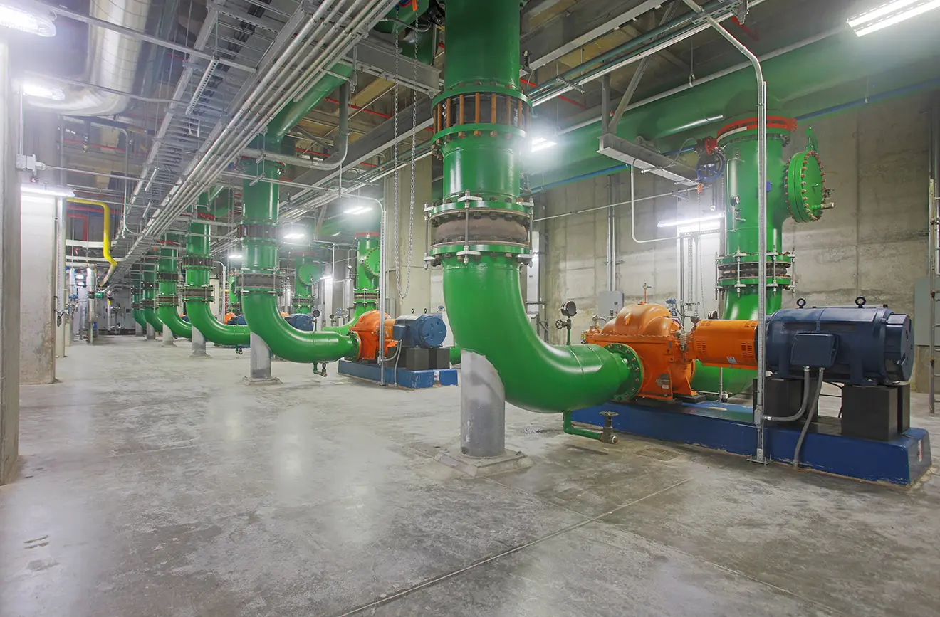 Spacious industrial room with large green pipes and orange machinery along the floor. Overhead pipes and bright lighting create a clean, orderly look.