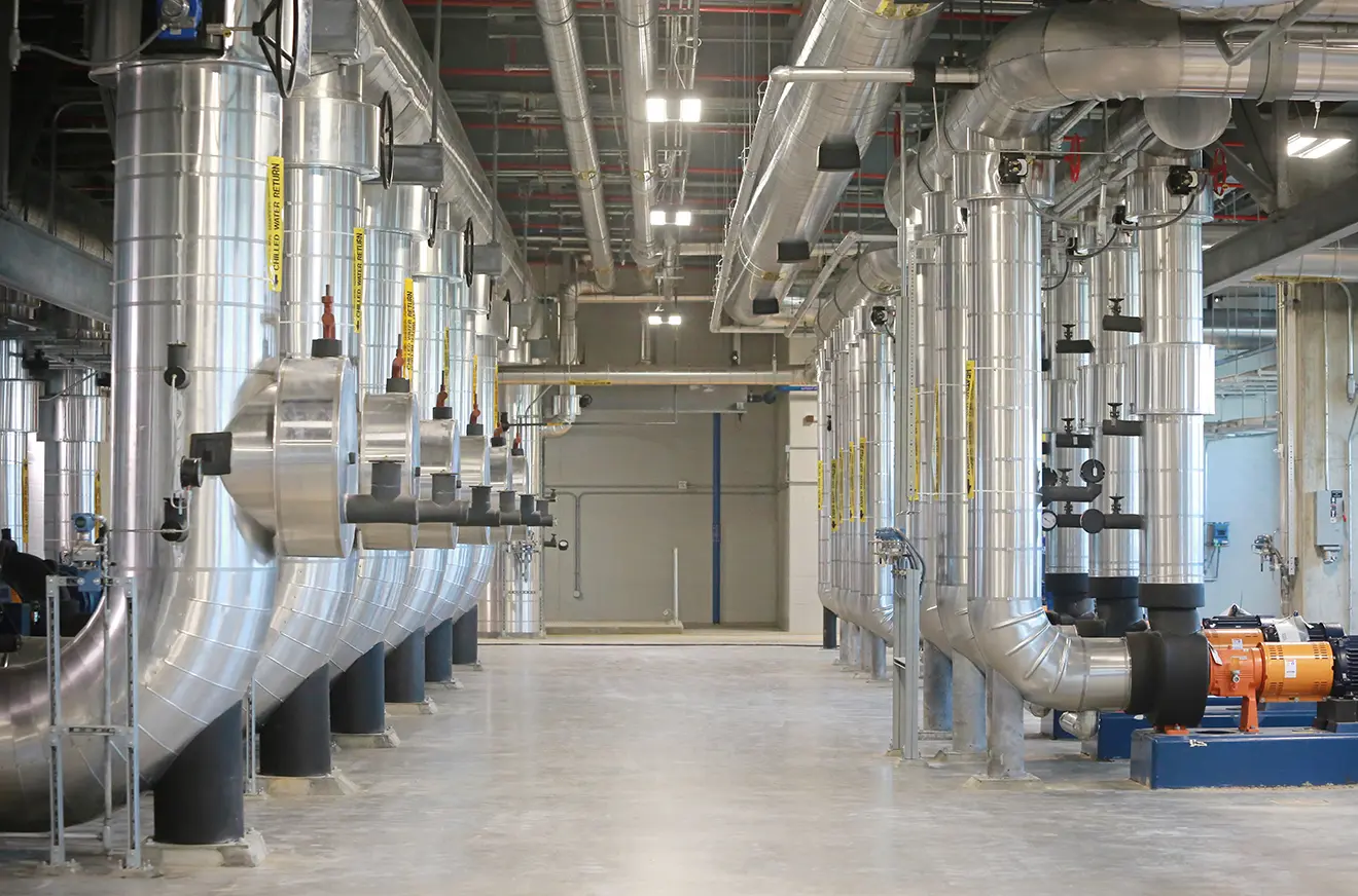 A large, brightly lit industrial room with rows of shiny metal pipes and ducts on both sides. The clean, organized space conveys a sense of efficiency.