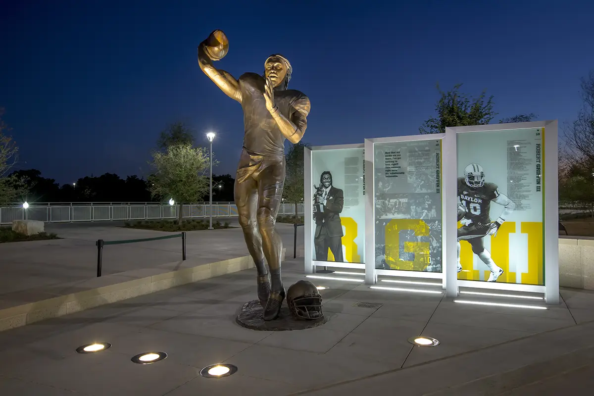 Bronze statue of a football player in a throwing pose, illuminated at night. Nearby are panels displaying images and text. A helmet rests at the statue's base.