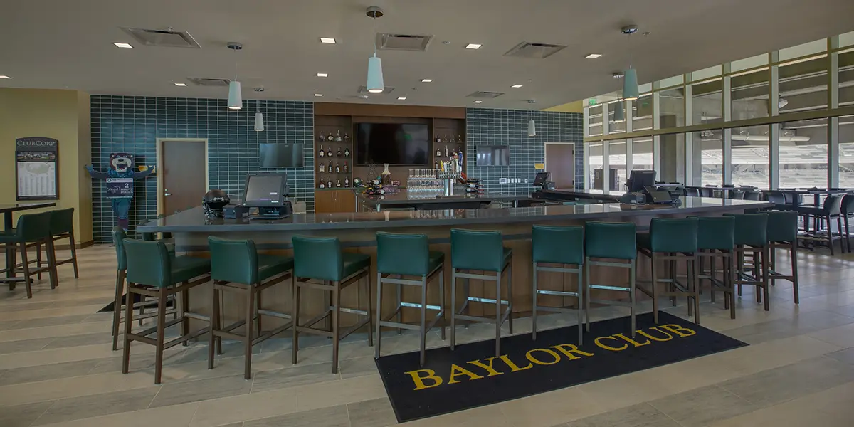 Modern bar interior with teal chairs and a central counter in a spacious room. A sign on the floor reads "Baylor Club." The atmosphere is sleek and inviting.