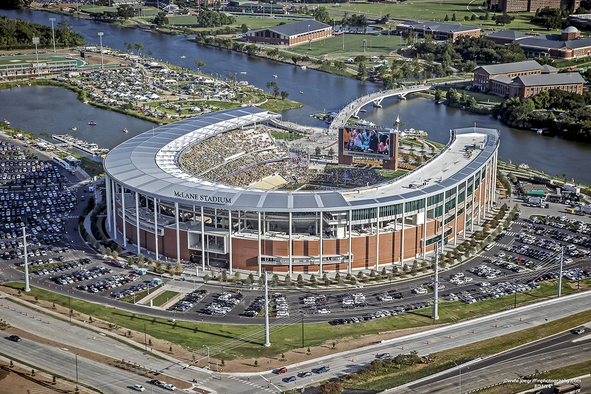 Aerial view of McLane Stadium, a large, oval sports venue beside a river. The stadium is packed with fans, surrounded by parking lots and greenery.