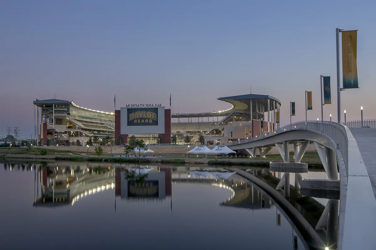 Stadium at dusk with glowing lights and a reflective river in the foreground. A modern bridge stretches over the water, leading to the entrance.