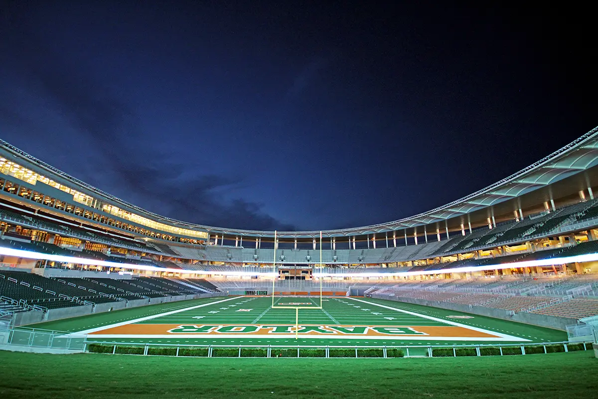Wide-angle view of an empty football stadium at dusk, featuring a brightly lit field with the word "Baylor," surrounded by tiered stands under a dark blue sky.