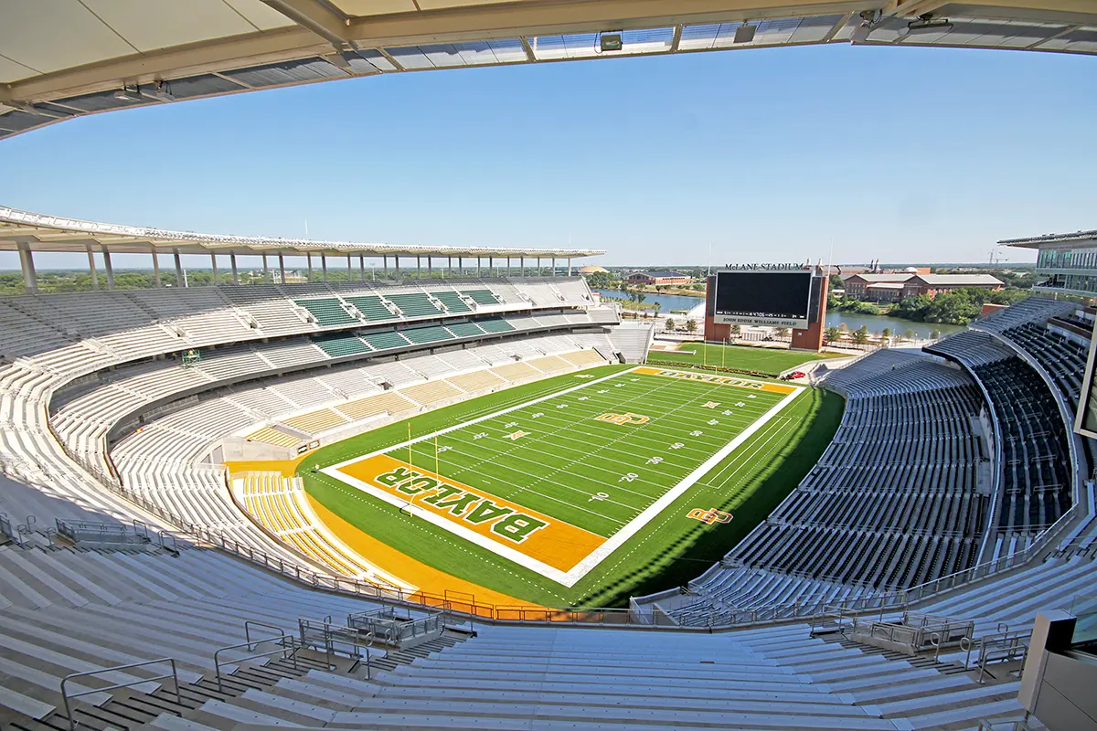 Aerial view of an empty football stadium with "Baylor" on the field. Bright green turf, expansive seating, large video screen, clear blue sky.