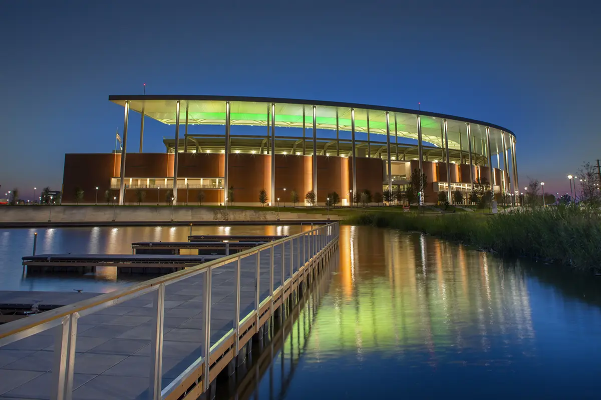 Aerial view of a large football stadium filled with spectators, vibrant green field, and prominent "Baylor Bears" signage. Surrounding parking lots and roads visible.