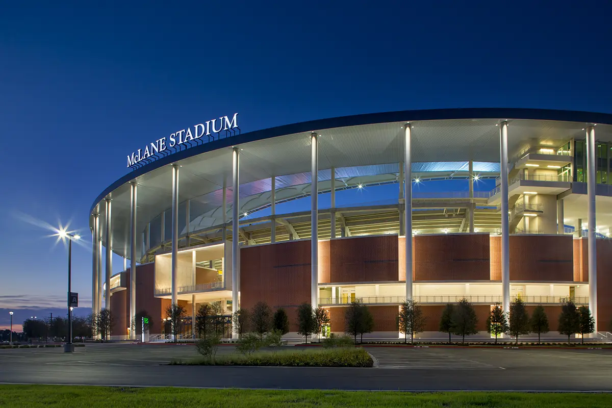 Modern stadium exterior at dusk with "McLane Stadium" sign illuminated. The structure is bathed in soft light under a deep blue sky, exuding a calm atmosphere.