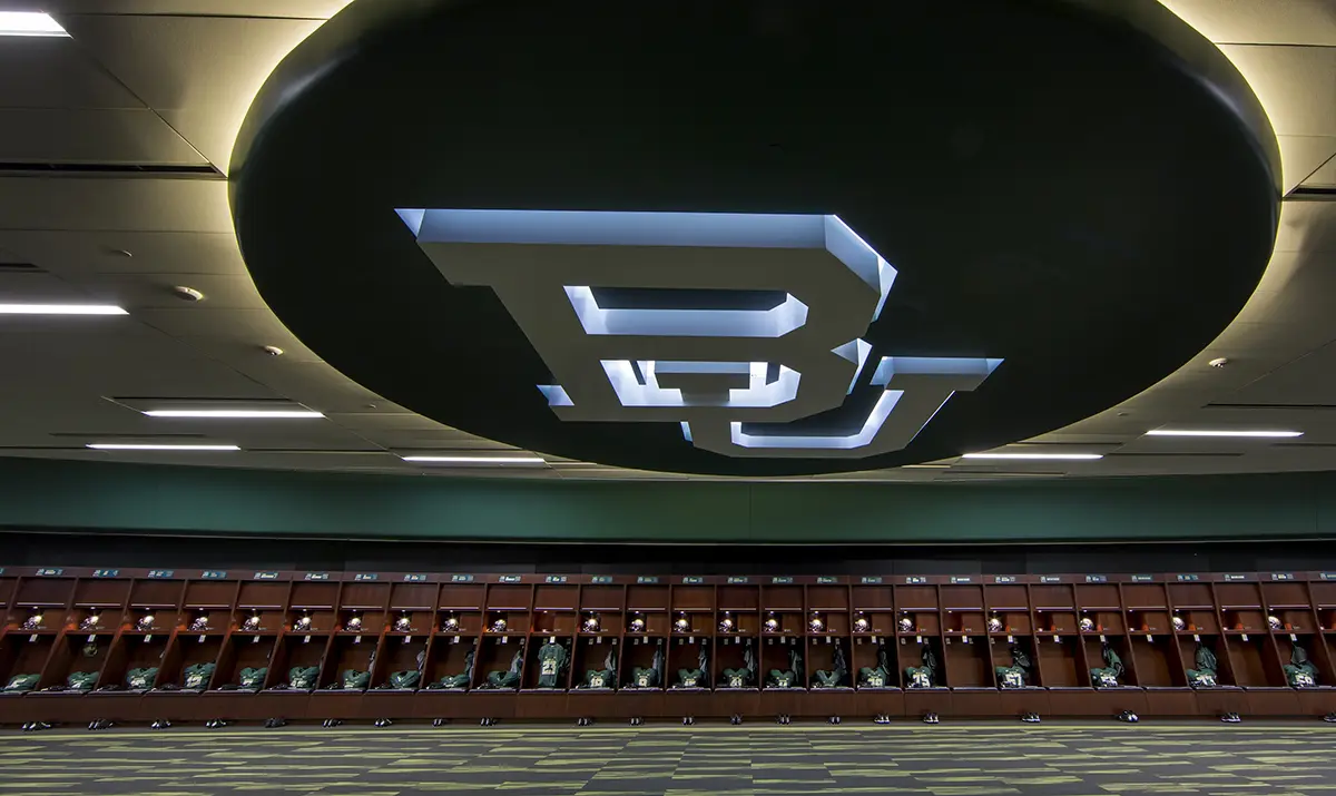 Aerial view of a large football stadium filled with spectators, vibrant green field, and prominent "Baylor Bears" signage. Surrounding parking lots and roads visible.