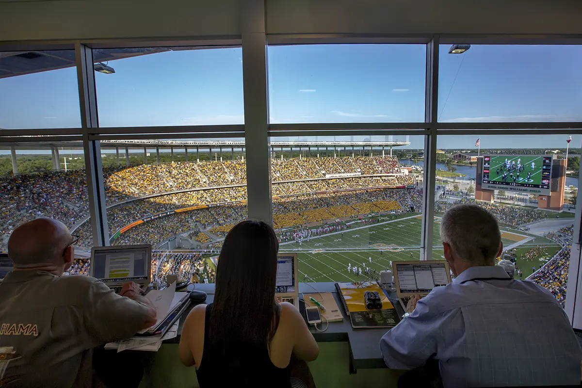 Three people view a sunny football game from a press box with laptops. A full stadium and field are visible, creating an energetic atmosphere.