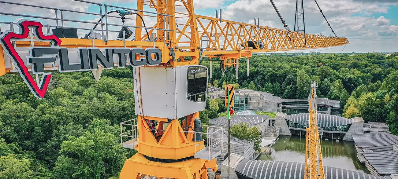 A large yellow crane labeled "Flintco" towers over a forested area and a modern building complex with curved roofs, under a partly cloudy sky.