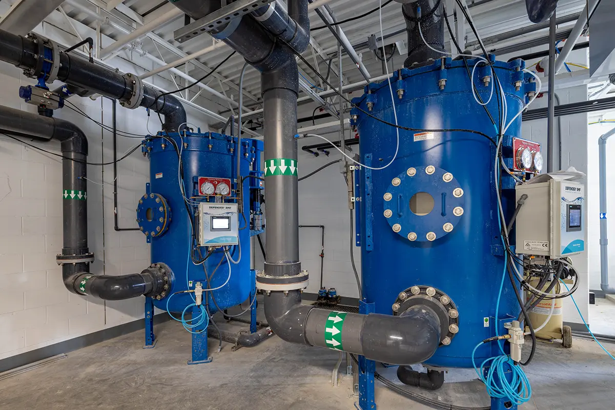 Industrial setting with large blue tanks connected by black pipes, featuring pressure gauges and control panels. The space is clean and well-organized.