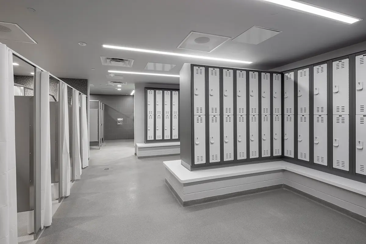 A modern locker room with rows of white lockers on the right, grey floor, and shower stalls with curtains on the left. Bright, clean, and organized.
