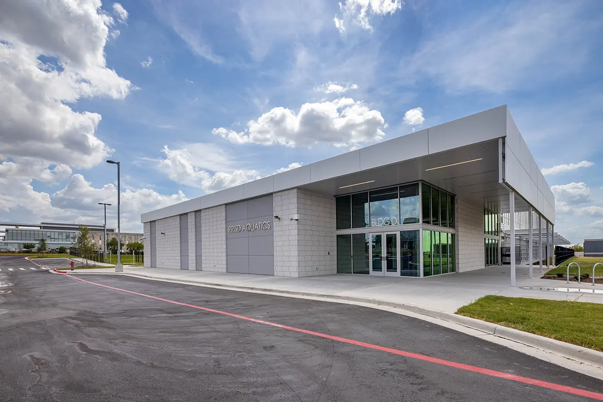 Modern building under a blue sky with scattered clouds. The structure features large glass windows, a flat roof, and a surrounding paved area.