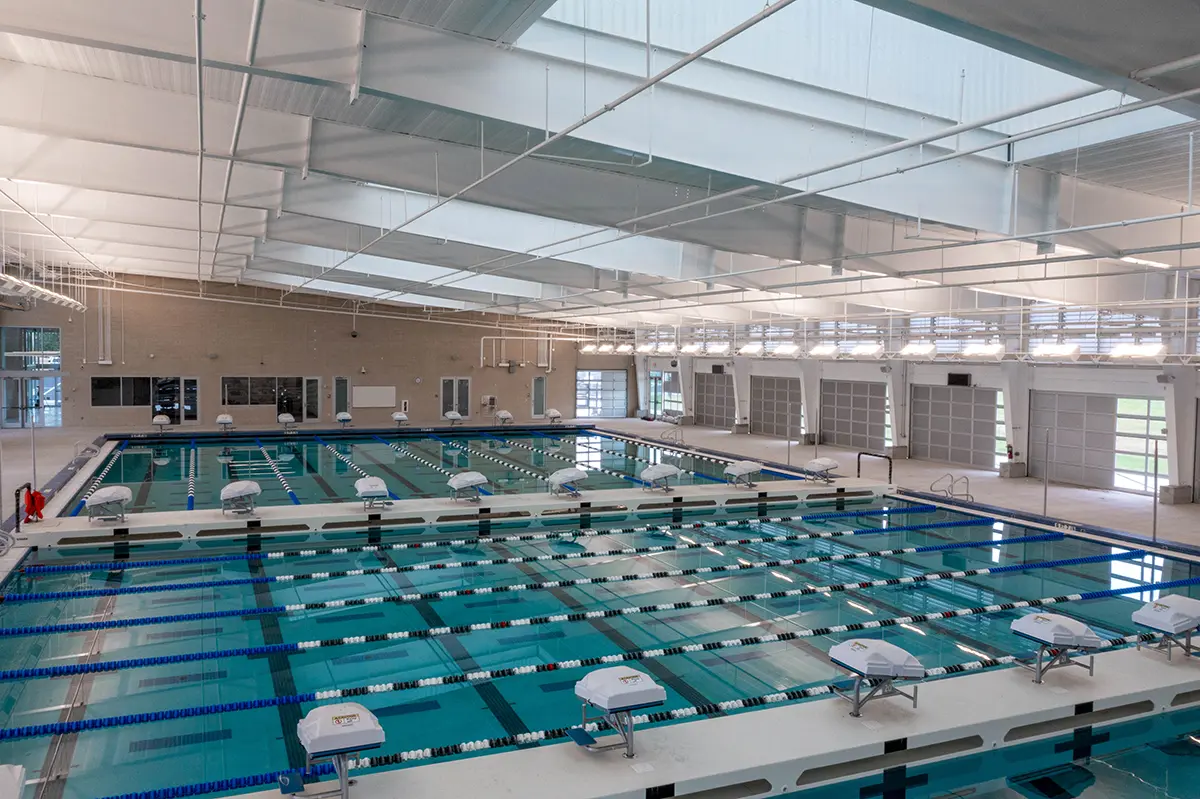 Spacious indoor swimming pool with clear lanes, starting blocks, and bright lighting. The modern facility has large windows and a high ceiling.