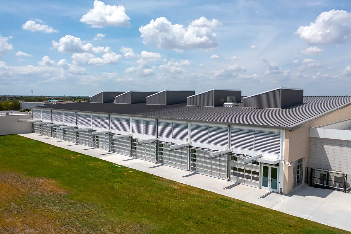 Modern industrial building with large glass-panelled doors and angular roof structures against a cloudy blue sky. The surrounding area has green grass, creating a serene and open atmosphere.