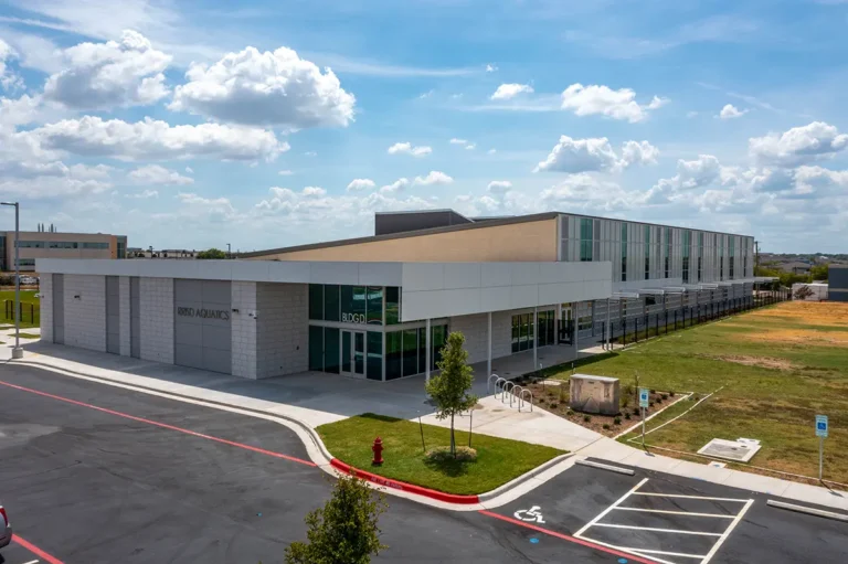 Modern school building under a bright blue sky with scattered clouds. The angular structure features large windows, surrounded by parking spaces and green lawns.