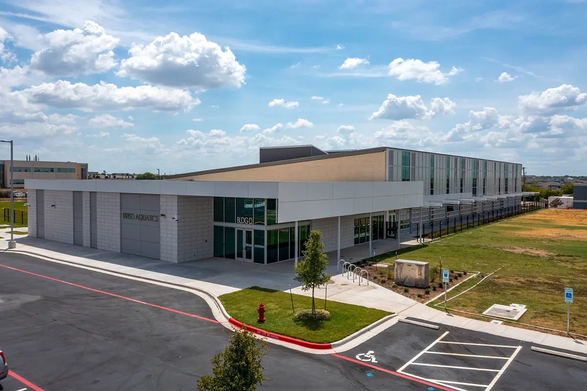 Modern school building under a bright blue sky with scattered clouds. The angular structure features large windows, surrounded by parking spaces and green lawns.