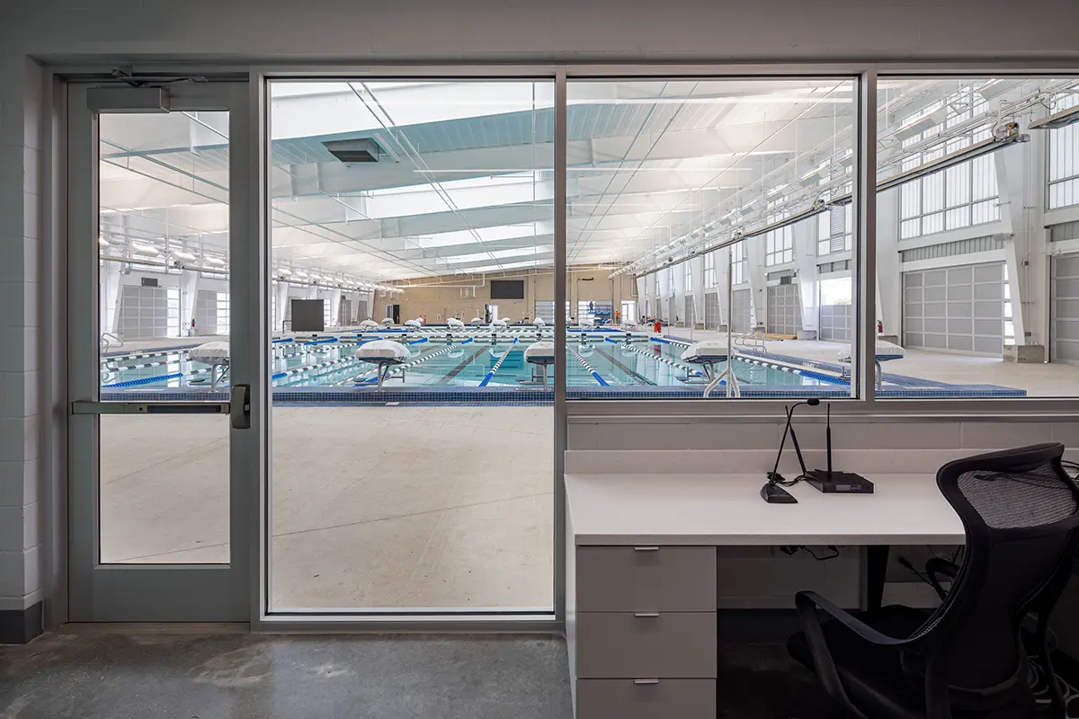 View from a modern office with a desk and chair, looking through glass windows at an indoor swimming pool facility, conveying a calm and clean atmosphere.