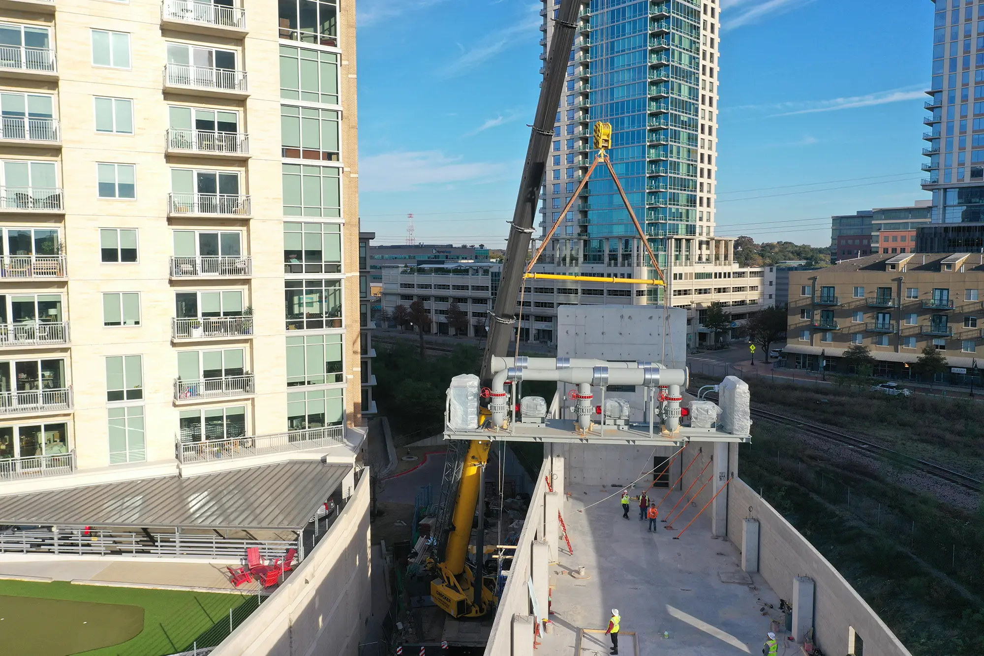 A large crane is hoisting a heavy piece of equipment between two tall buildings in an urban setting. Workers in safety gear are guiding the process from below. The scene conveys a tone of precision and teamwork.