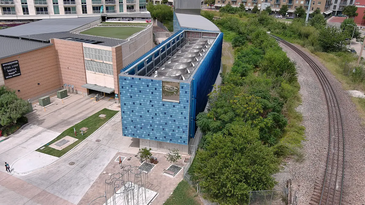 Aerial view of a modern building with a blue tiled facade next to railroad tracks and greenery. People walk near it, creating a peaceful urban scene.