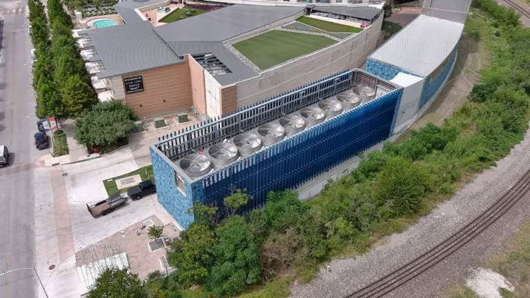 Aerial view of a modern industrial building with blue and beige tones, surrounded by greenery. Rooftop features multiple industrial fans. Nearby, a road and train tracks.