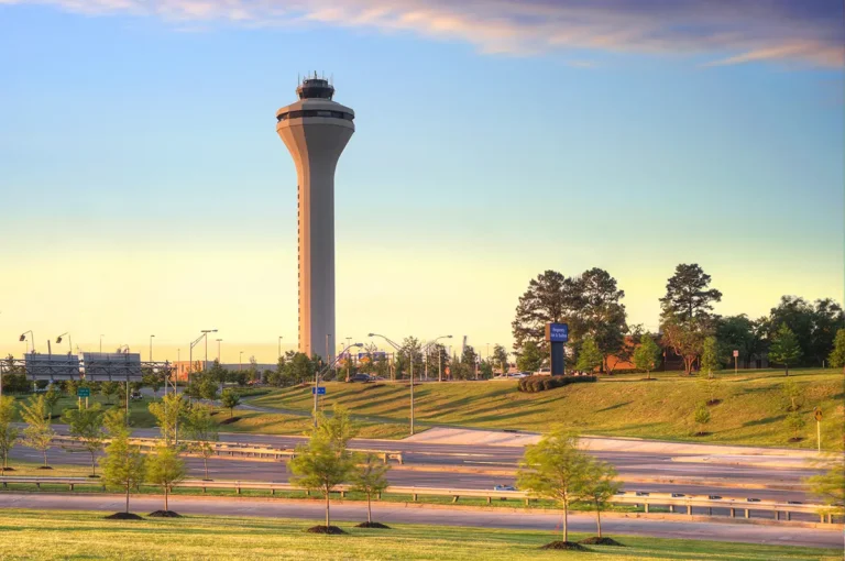 A tall airport control tower stands against a serene, sunset-lit sky. Surrounded by green lawns and trees, the scene conveys a peaceful, evening atmosphere.