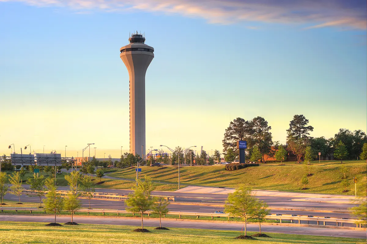 A tall airport control tower stands against a serene, sunset-lit sky. Surrounded by green lawns and trees, the scene conveys a peaceful, evening atmosphere.