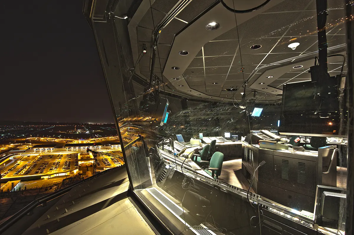 Nighttime airport control tower interior with dim lighting, showing empty chairs, computer monitors, and a panoramic view of runway lights outside.