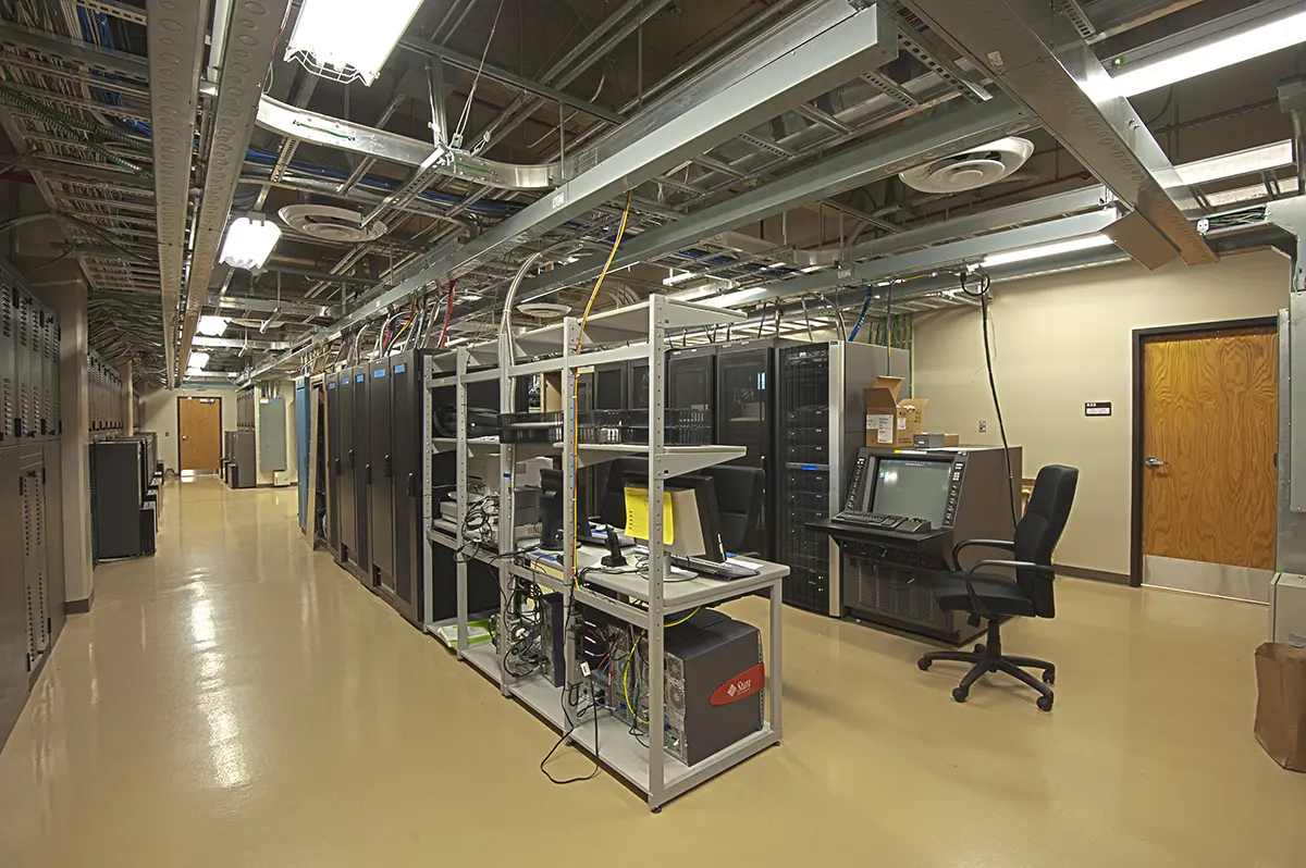 A spacious server room with numerous black server racks lined up. Overhead cables and bright lights are visible. A chair and monitor stand in the corner.