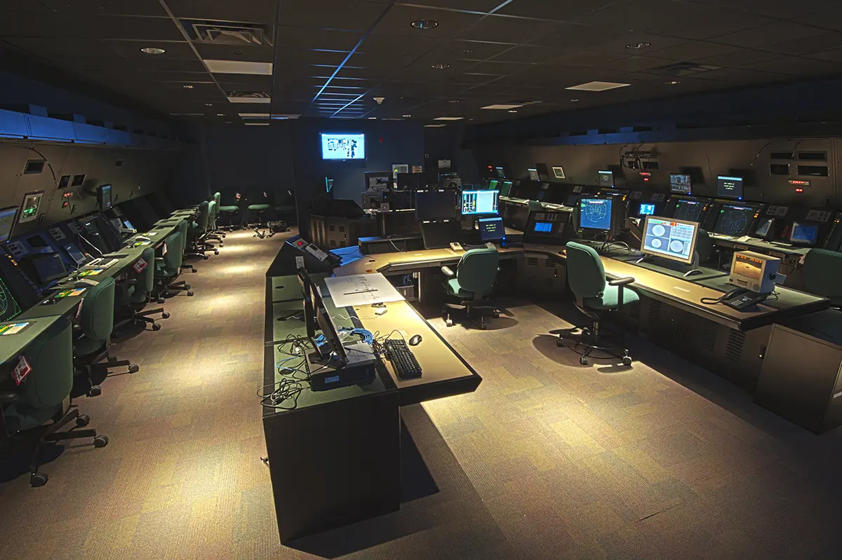 Dimly lit control room with multiple computer stations, monitors, and green chairs arranged in a U-shape; an atmosphere of focus and technology.