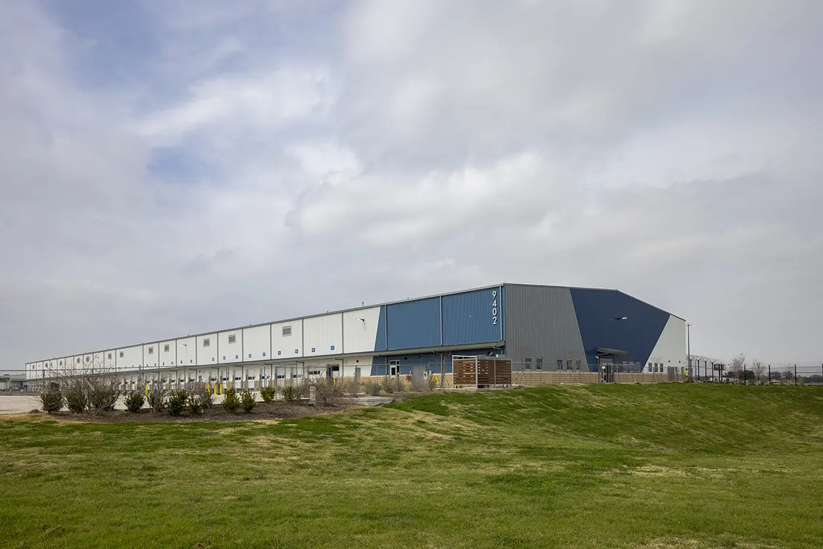 Large industrial warehouse with blue and white panels, surrounded by green grass, under a partly cloudy sky. The scene feels expansive and orderly.