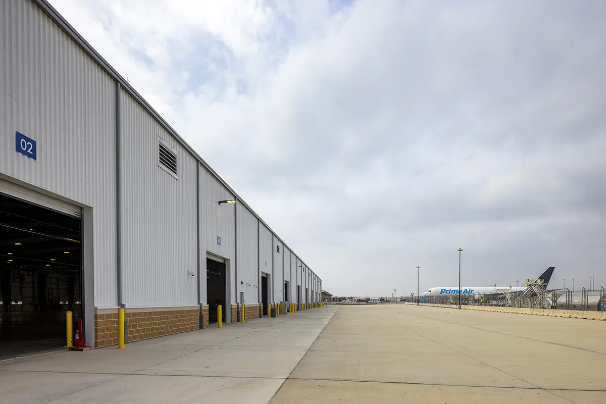 Industrial warehouse exterior on a cloudy day, featuring an open garage, yellow safety bollards, and a distant airplane on the tarmac.