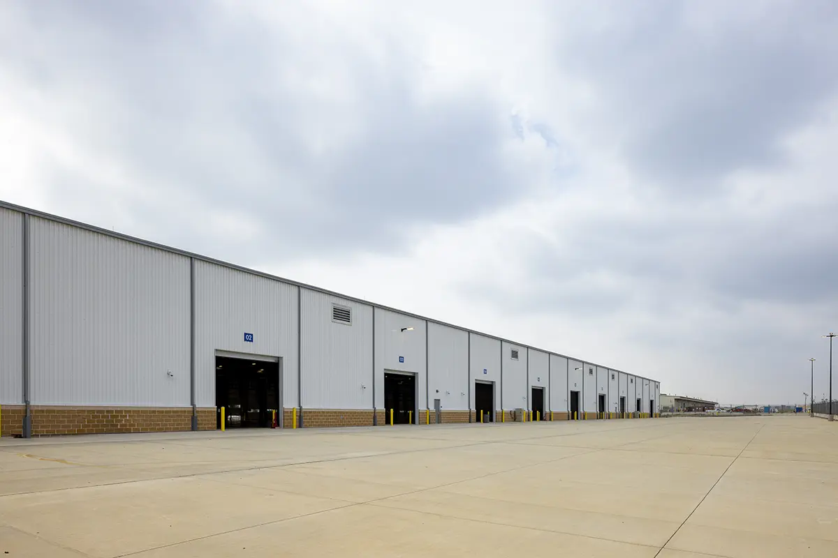 A long industrial warehouse with multiple loading bays under a cloudy sky. The building's exterior is light gray and brown, conveying a sense of spaciousness.