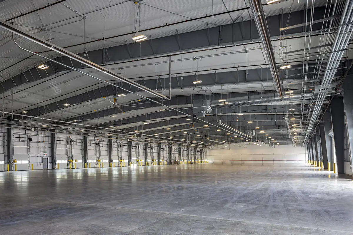Spacious, empty warehouse interior with high ceilings, steel beams, and multiple loading docks on the left. Bright overhead lighting creates a clean, industrial feel.