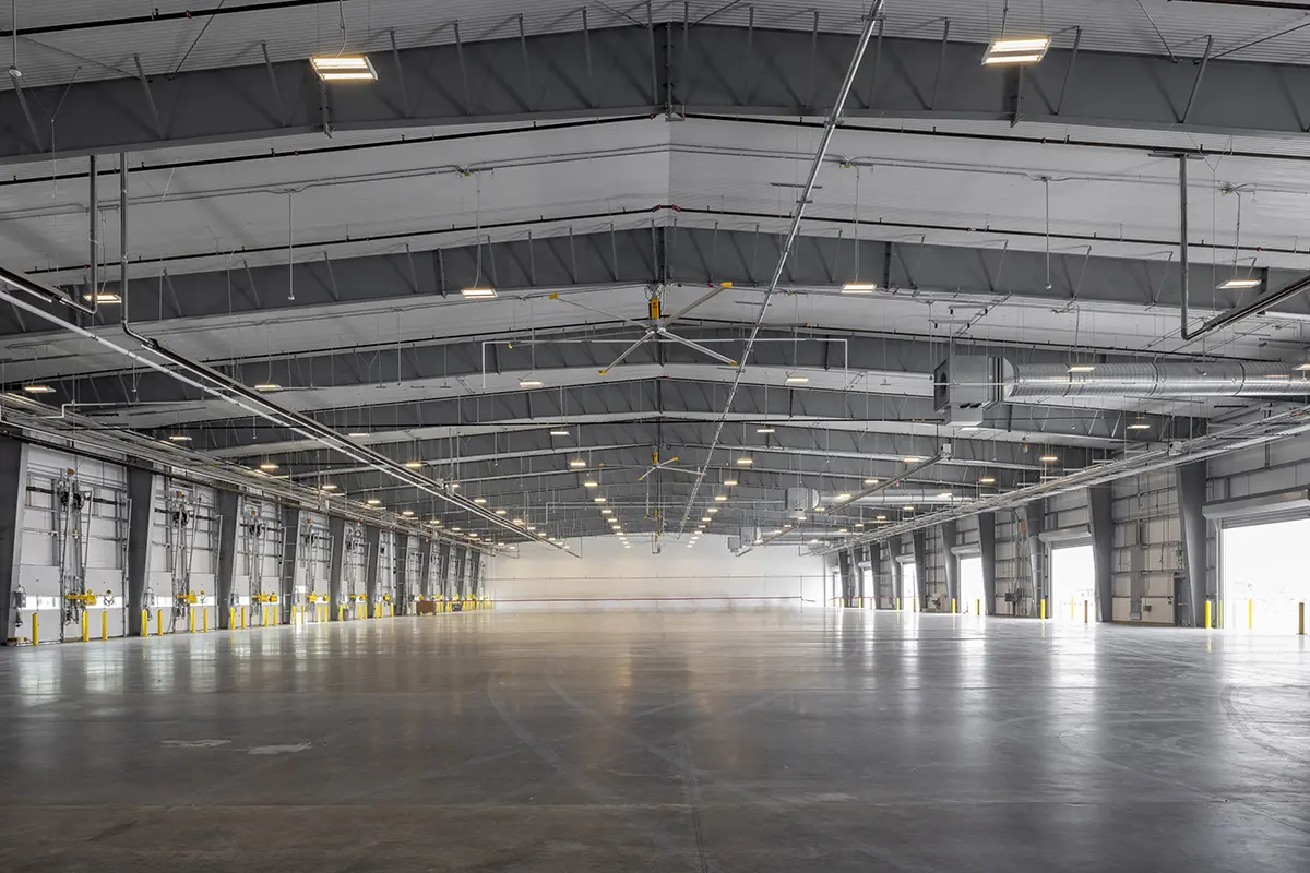 Expansive, empty warehouse interior with a high, arched ceiling. Industrial lighting and pipes run along the ceiling, creating a vast, open space.