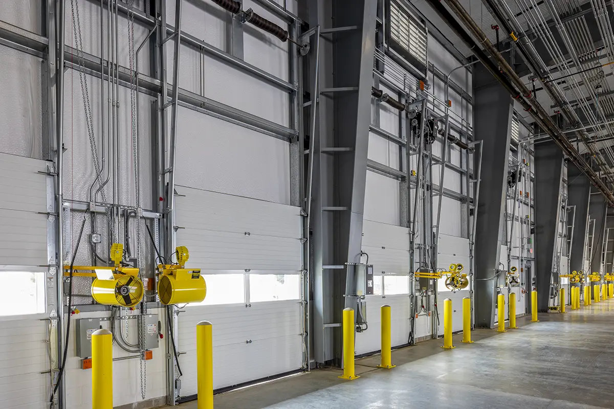 Inside a spacious warehouse with large loading bay doors and bright yellow safety bollards. Overhead pipes and cables add an industrial feel.