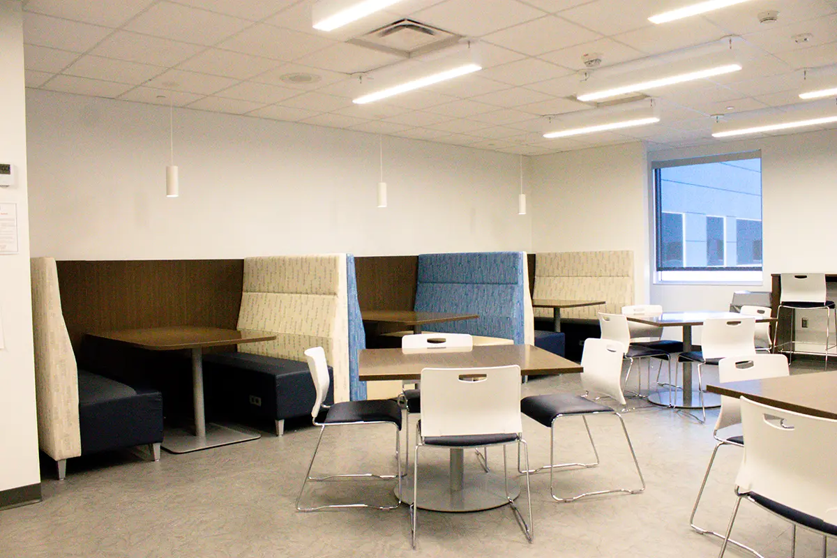 Modern cafeteria with white walls, square tables, white and blue chairs. Cozy booth seating lines the wall. Large windows provide natural light.