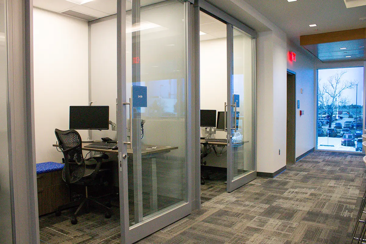 A modern office hallway with glass-walled meeting rooms, each with a computer, desk, and chair. The space appears clean and professional.