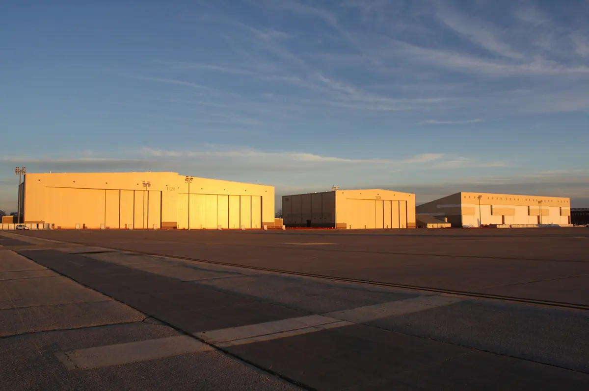 Three large airplane hangars bathed in golden sunlight on an expansive tarmac, set against a blue sky with wispy clouds, conveying calmness.