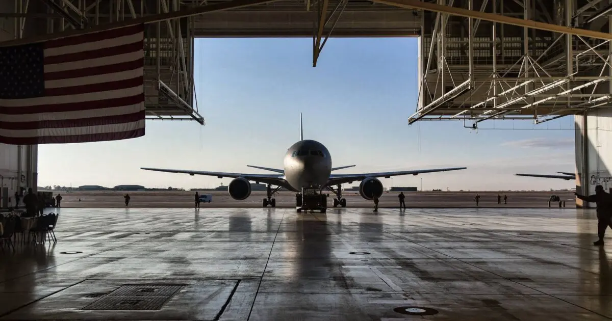 Airplane in a hangar, viewed from inside with an American flag hanging on the left. The open hangar reveals a sunny runway and clear blue sky.